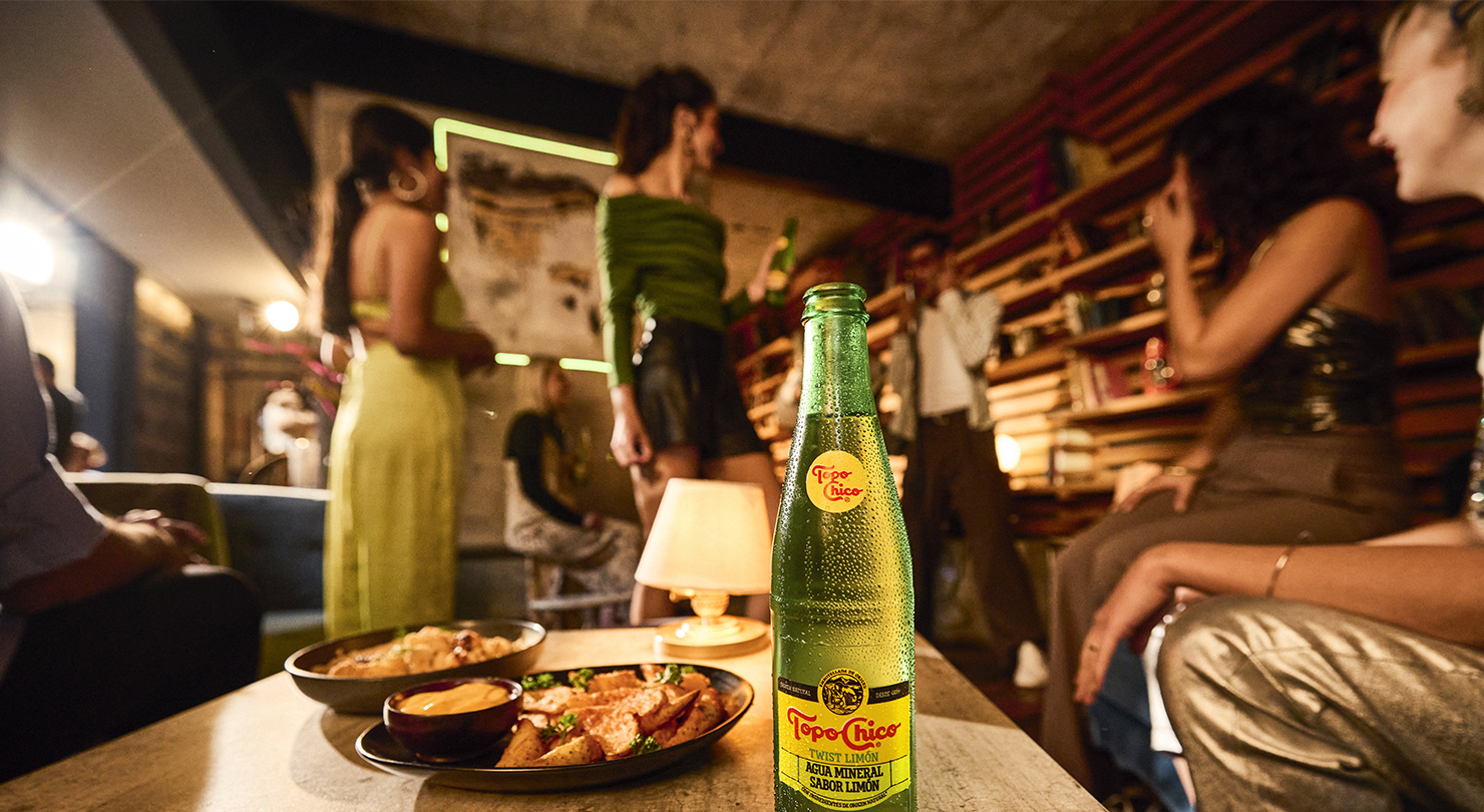 Una botella de Topo Chico Twist Limón, está en primer plano sobre una mesa con bocadillos y un grupo desenfocado de personas hablando en el fondo.