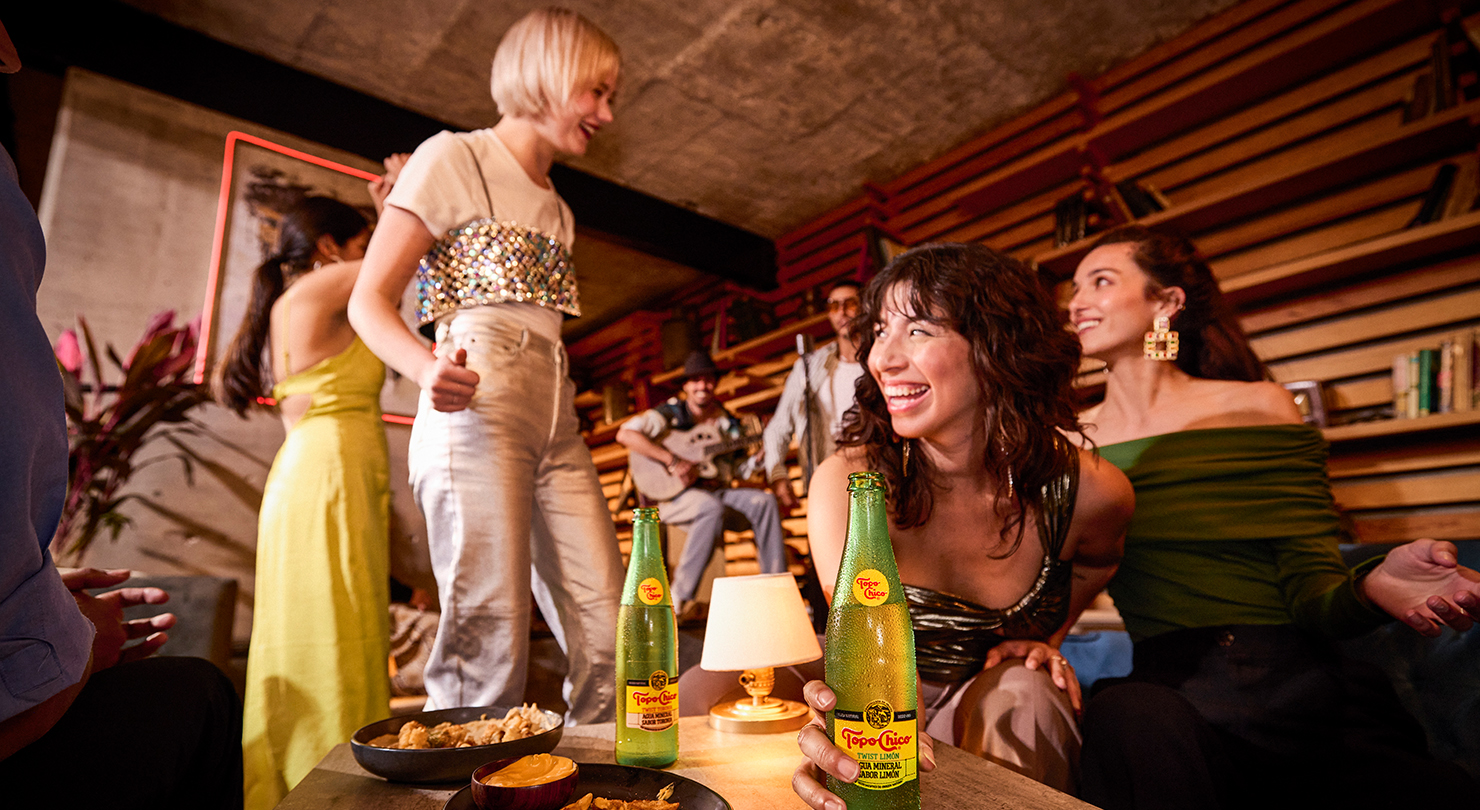 Amigos reunidos en una fiesta con Topo Chico y bocadillos, con un músico tocando al fondo.