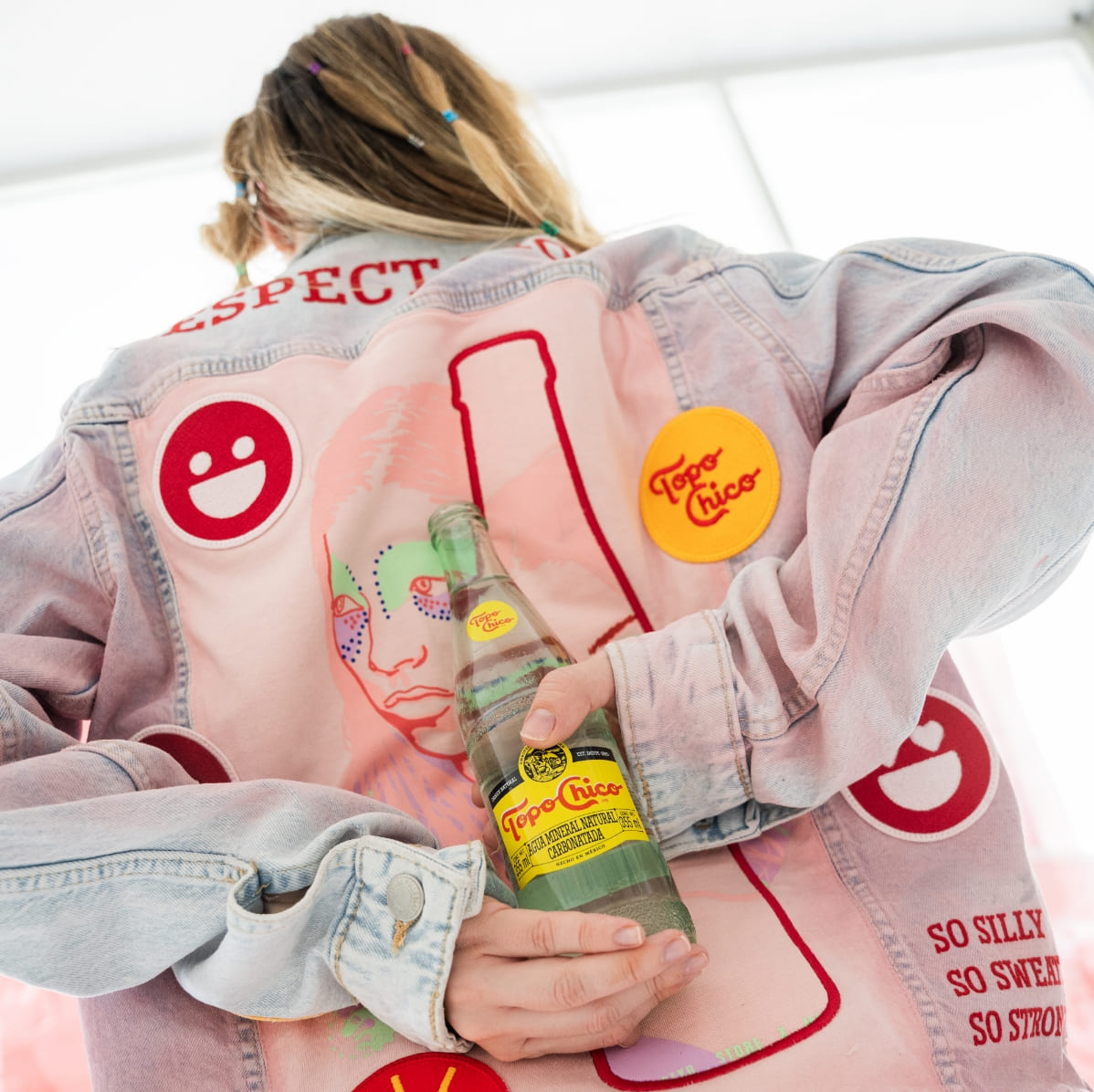 Una chica de espaldas con una Jean Jacket estampado de colores rosa y rojo y está sosteniendo una botella de vidrio Topo Chico